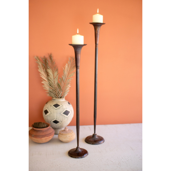 Two Vail Metal Candle Holders with an antique copper finish, topped with lit white candles, form a striking centerpiece. Behind them, pottery vases and dried palm leaves stand out against a muted orange wall for stylish mantle decor.