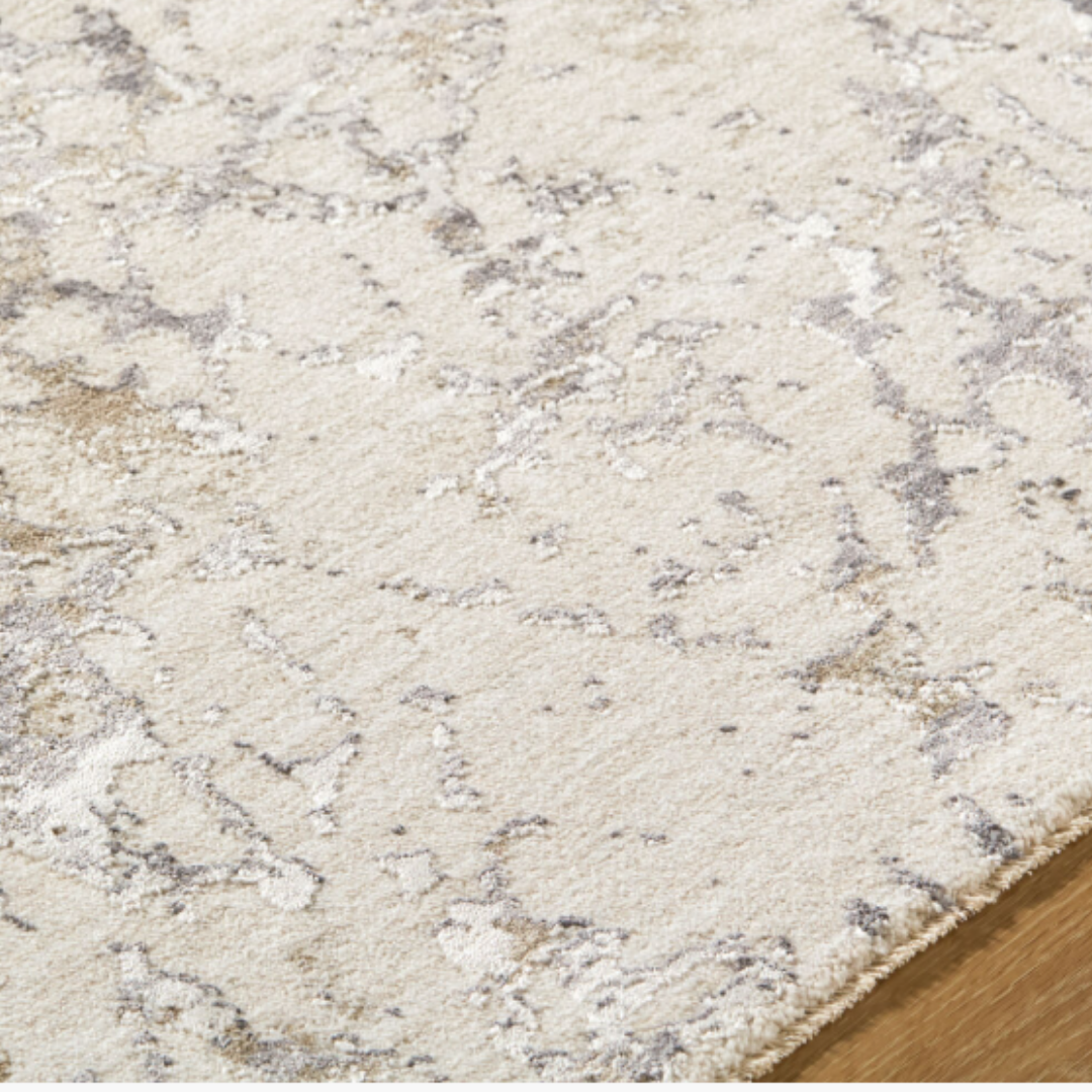 A close-up of the Velmira Machine Woven Rug reveals its soft beige and light grey texture with an abstract marbled pattern. Cream and taupe tones highlight its modern craftsmanship and durability on a wooden floor.
