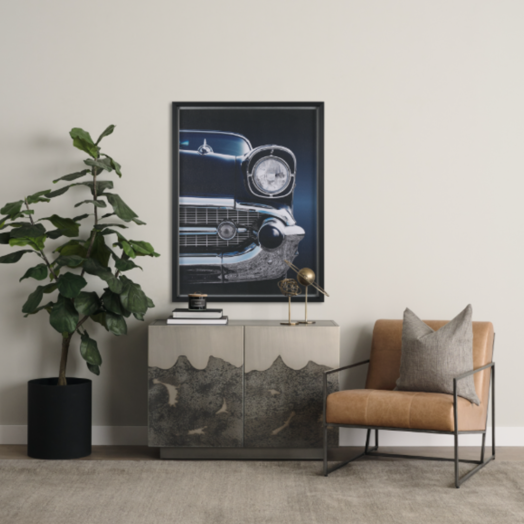 In the stylish living room, a potted plant sits on the left, while a modern distressed cabinet centers the space. A classic car print hangs above, and on the right, a Vercelli Handcrafted Leather Accent Chair enhances the mid-century modern vibe with minimalist neutral tones.
