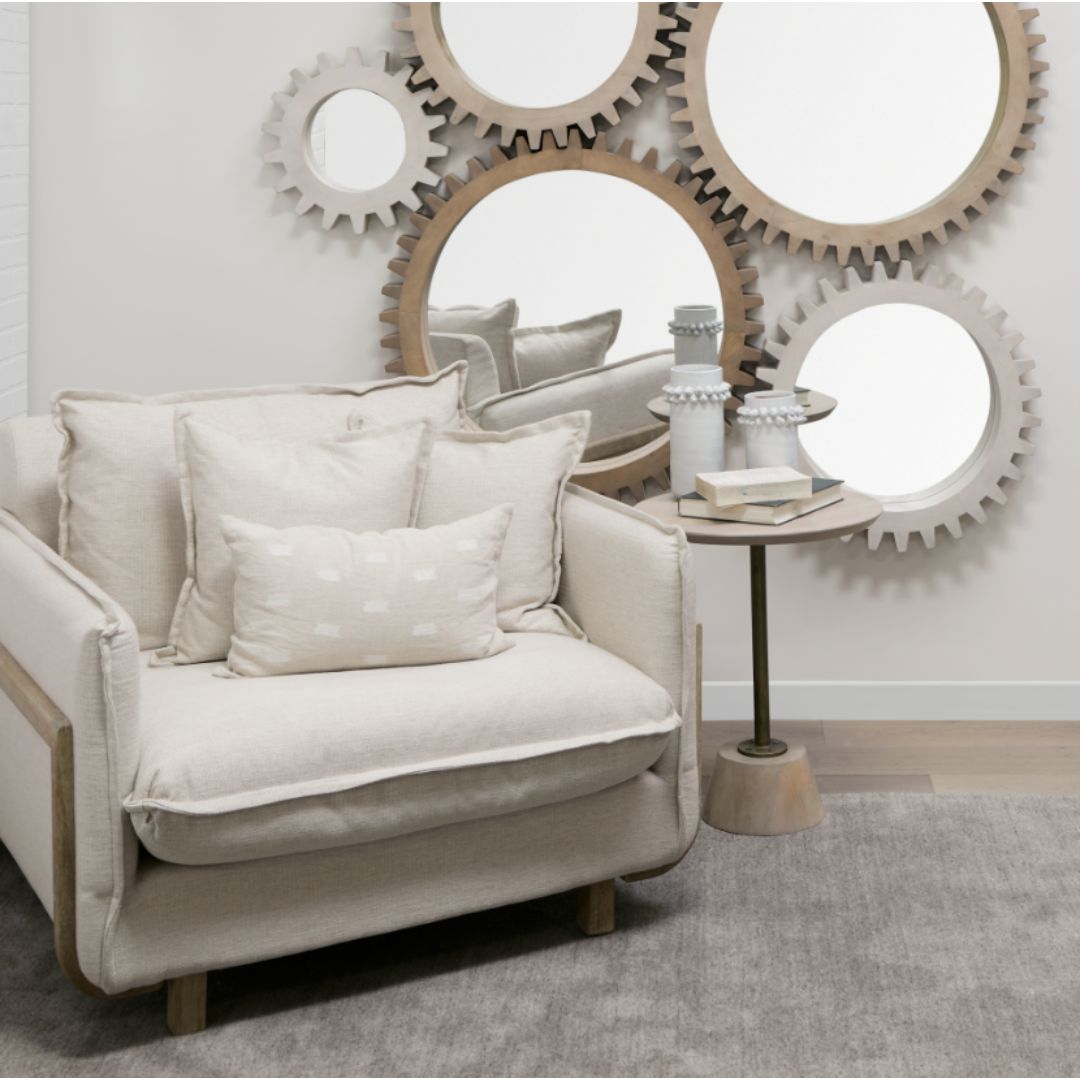 A cozy room features the Veyra Plush Upholstered Armchair in beige, offering plush comfort with two cushions. A side table holds books and ceramic vases, while interlocking gear-shaped mirrors enhance the minimalist aesthetic. A soft neutral rug adorns the solid oak floor.