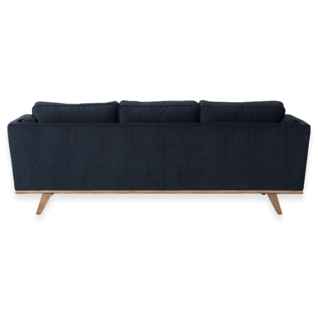 The Zinnia Mid-Century Modern Navy Fabric Sofa is shown from the back with three reversible cushions, angled wooden legs, and a light wood trim along the bottom edge, set against a white background.