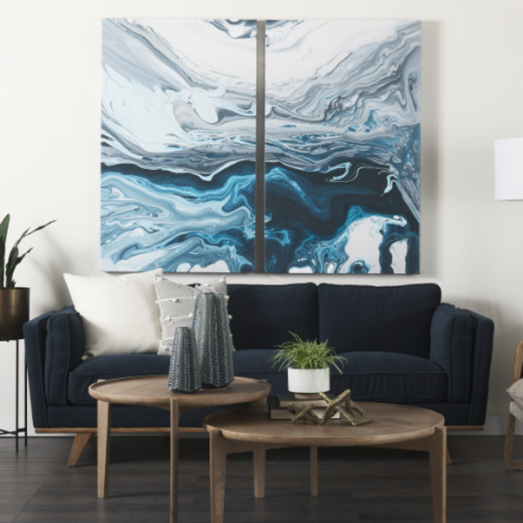A modern living room highlights the Zinnia Mid-Century Modern Navy Fabric Sofa with white and gray reversible cushions, paired with round wooden coffee tables, vases, plants, and abstract blue wall art for a stylish look.