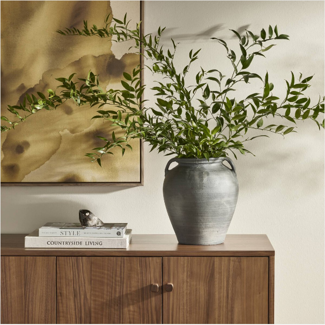 A Black Faith Terracotta Vase filled with lush green branches sits atop a wooden cabinet. Next to it, two stacked books titled "Style" and "Countryside Living" support a decorative piece: a small bird. A watercolor abstract painting with earthy tones hangs on the wall in the background, complementing the decor.