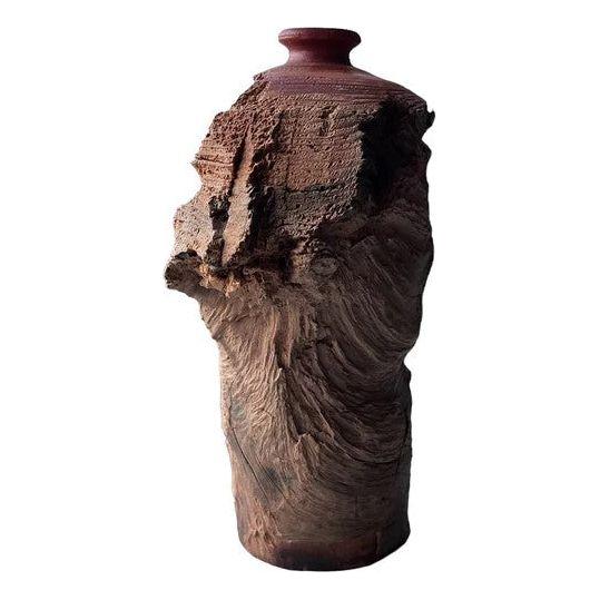 A tall, cylindrical ceramic vase with an unfinished, rugged texture on one side, giving it a natural, eroded appearance. The smooth upper portion and lip are contrasted by the rough, jagged surface below, creating a striking juxtaposition between refined and raw materials. This hand-carved piece from the Cloud Vase collection symbolizes prosperity through its unique design.