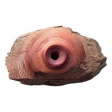 A piece of wood, likely driftwood, is shown with a smooth, rounded, hollow center. The wood displays rich, reddish-brown tones and intricate grain patterns radiating outward from the central hole. The edges are rough and textured, giving it a natural, weathered appearance reminiscent of hand-carved artisanship.