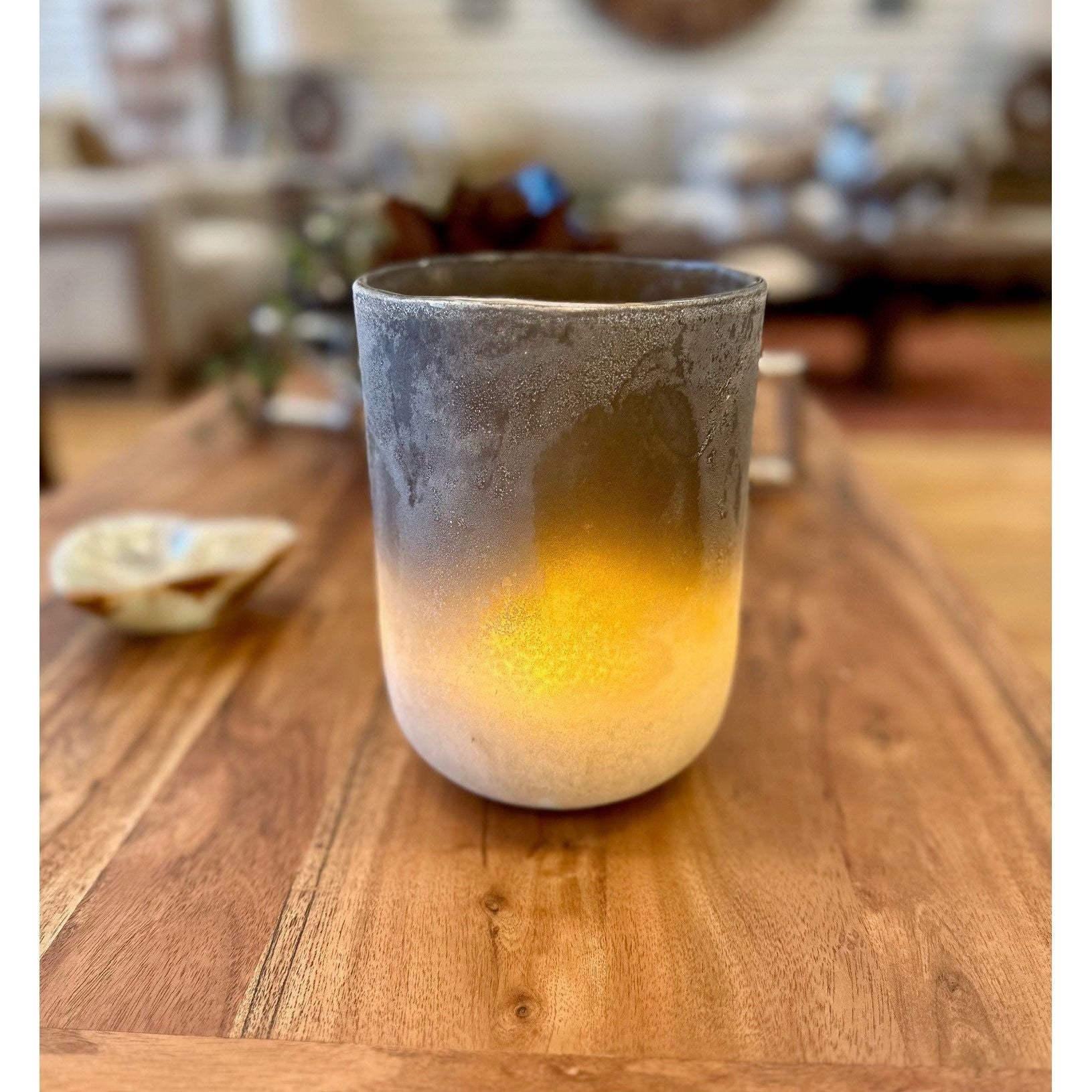 The Desert Sands Hurricane Vase, with its frosted, gradient glass holder, sits elegantly on a wooden table. The vase transitions from dark gray at the top to a warm yellow glow near the base, evoking earthy hues. The softly focused background displays blurred furniture and decorations, contributing to a cozy ambiance.