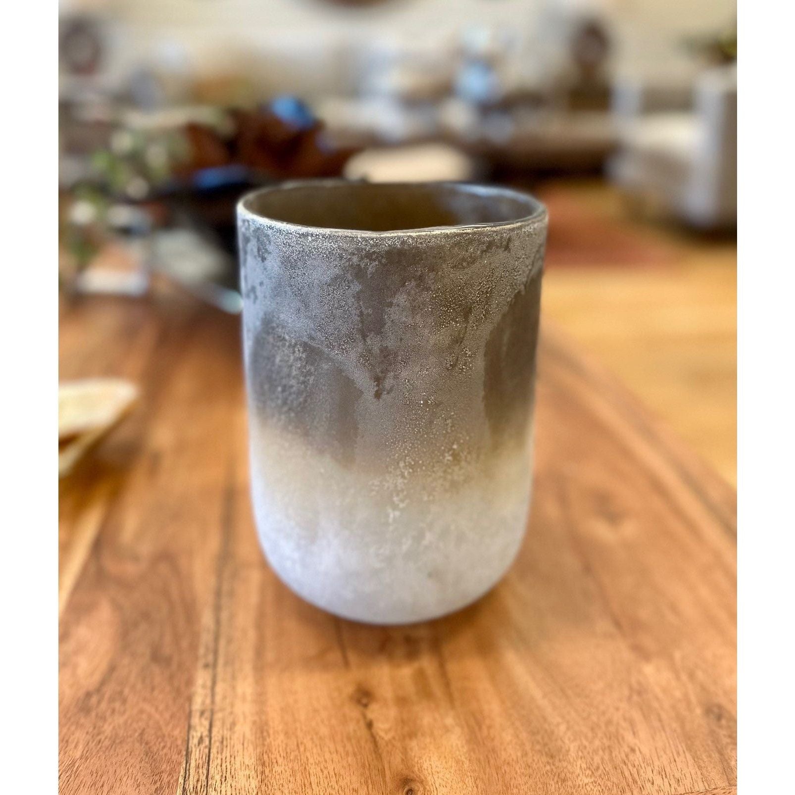 The Desert Sands Hurricane Vase with a matte finish is displayed on a wooden surface. The vase features a gradient color pattern, transitioning from darker grey at the top to light grey and almost white at the bottom, resembling earthy hues. The background is softly blurred, indicating a cozy indoor setting.