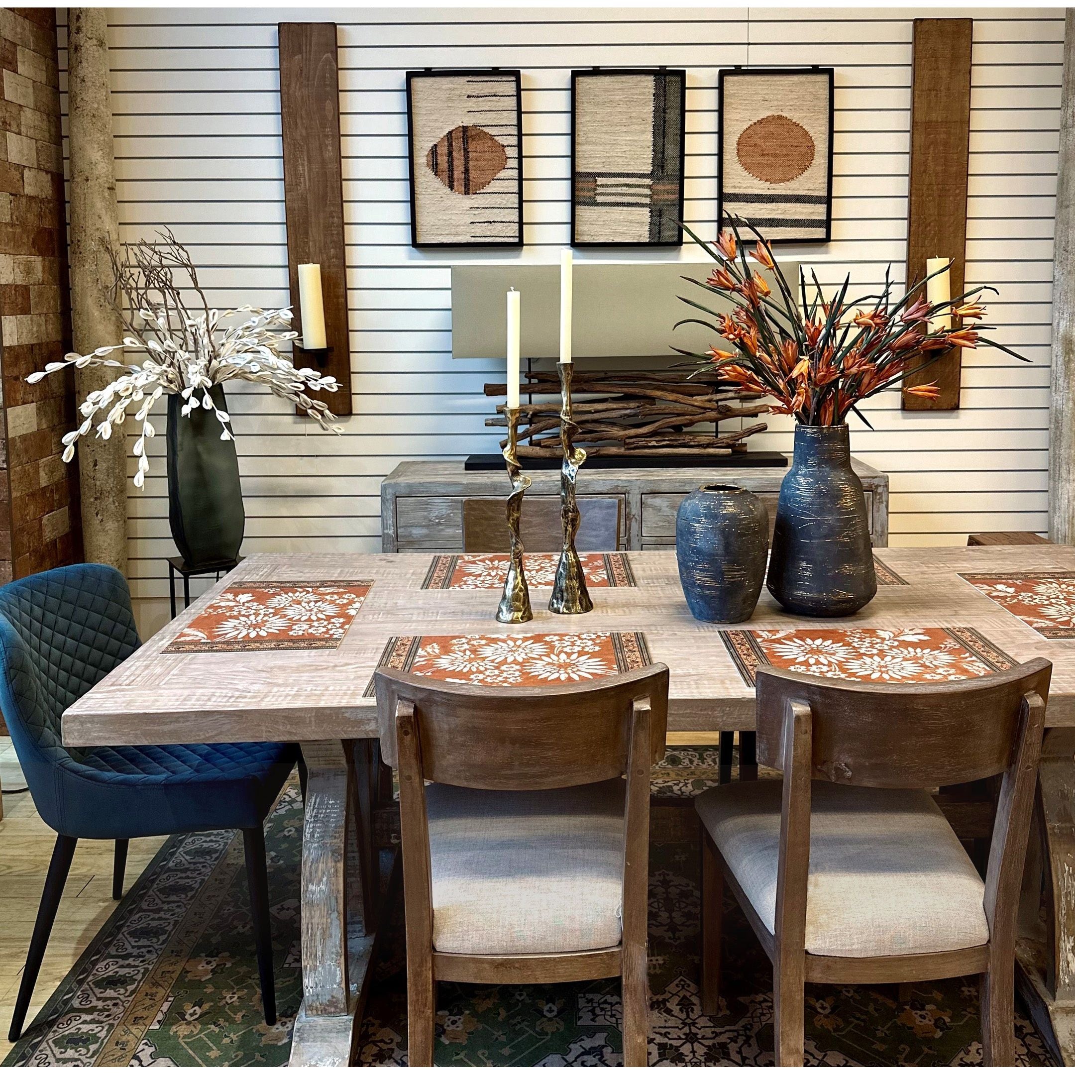 In a chic dining room, a solid elm table is dressed with patterned orange placemats and two tall white candles. It is surrounded by four chairs, including the Molly Dining Chair in medium grey/sand upholstery. The decor includes decorative wall panels and a sideboard with vases and plants.