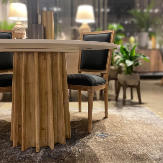 A round wooden dining table with a channeled oak wood base, named the Channel Concrete Dining Table, is surrounded by two chairs featuring black cushions and wooden frames. In the background, there are blurred out plants, lamps, and additional furniture, creating a cozy indoor setting. The floor is covered with a textured area rug.
