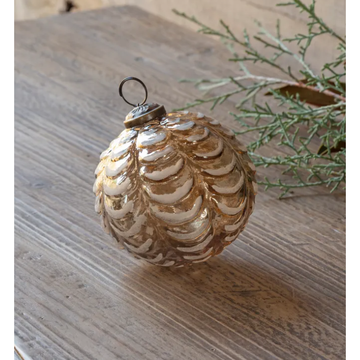 The Draped Frozen Glass Ornament, designed in the shape of a pine cone with vintage appeal, sits elegantly on a wooden surface. It boasts a metallic finish embellished with gold and white accents and features layered, scalloped textures. In the background, a sprig of greenery enhances the scene.