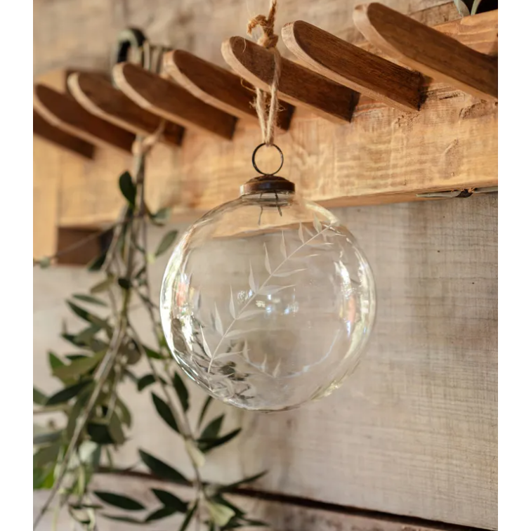 An Etched Glass Ornament with intricate leaf engravings is suspended from a wooden hook on a rustic wall. The natural wood and subtle wall tones enhance the greenery, evoking a tranquil, earthy atmosphere. The ornament's transparent surface gracefully reflects light and its surroundings.