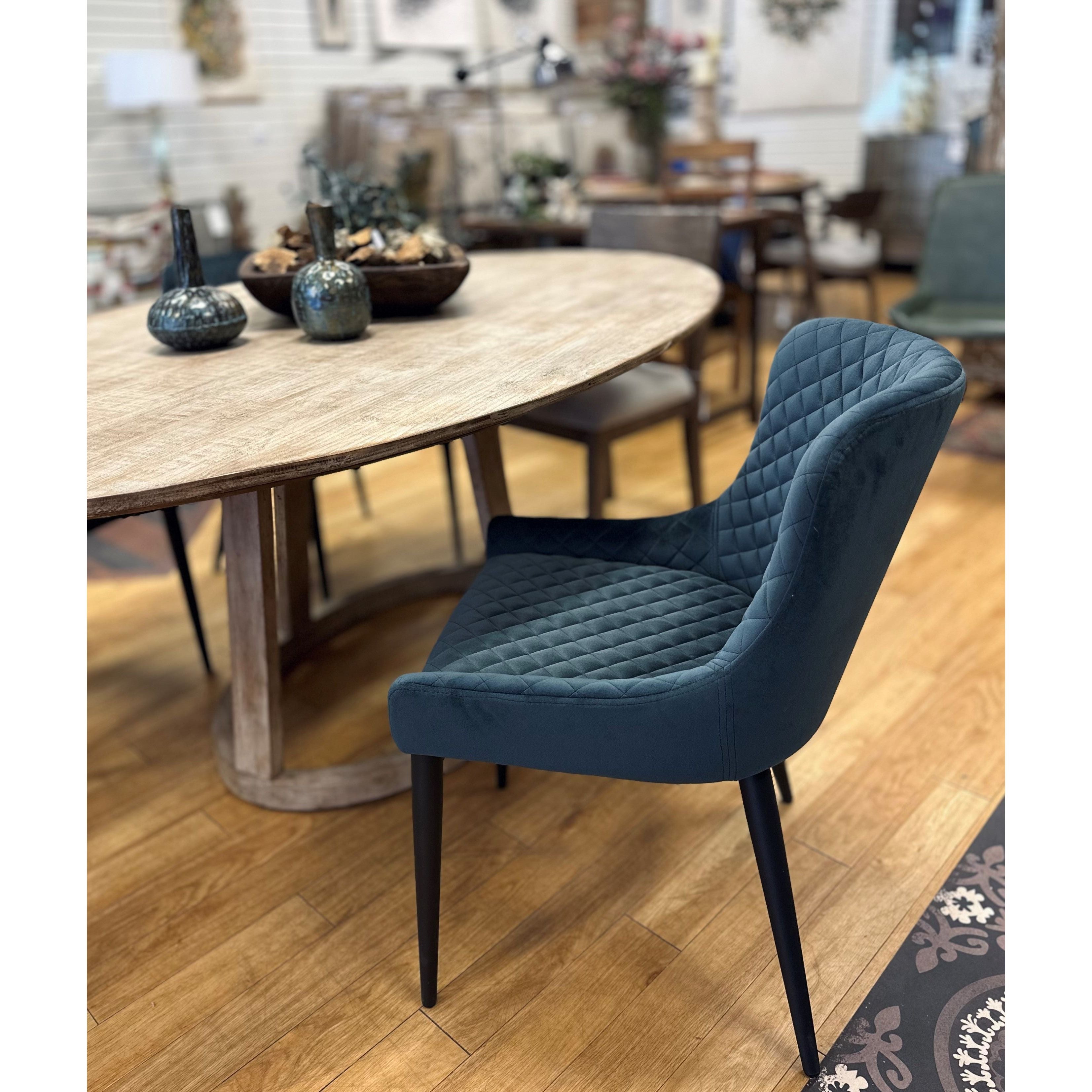 A cozy, well-furnished room featuring a large oval wooden table with a tufted treasure—an Etna Dining Chair in quilted dark teal with long black legs—in the foreground. On the table are decorative vases, small plants, and wooden bowls. The background has additional chairs, shelves, and wall decorations. Wood flooring.