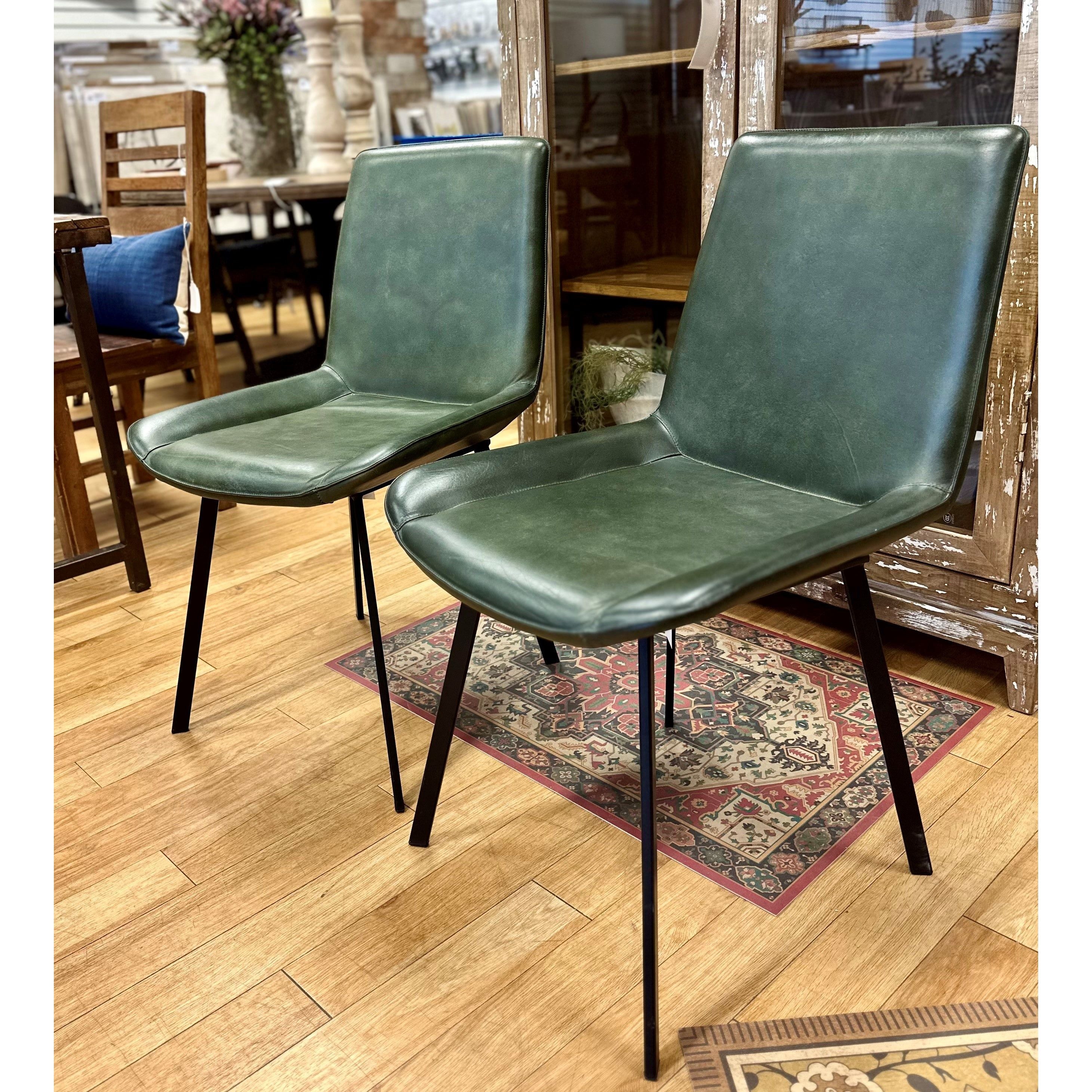 Two Gabby Dining Chairs in hunter green goat leather with black iron frames are placed together on a wooden floor. Behind them is a wooden cabinet with glass doors and a mirror reflecting parts of the room. A red patterned rug lies beneath the chairs, and various pieces of furniture populate the background.