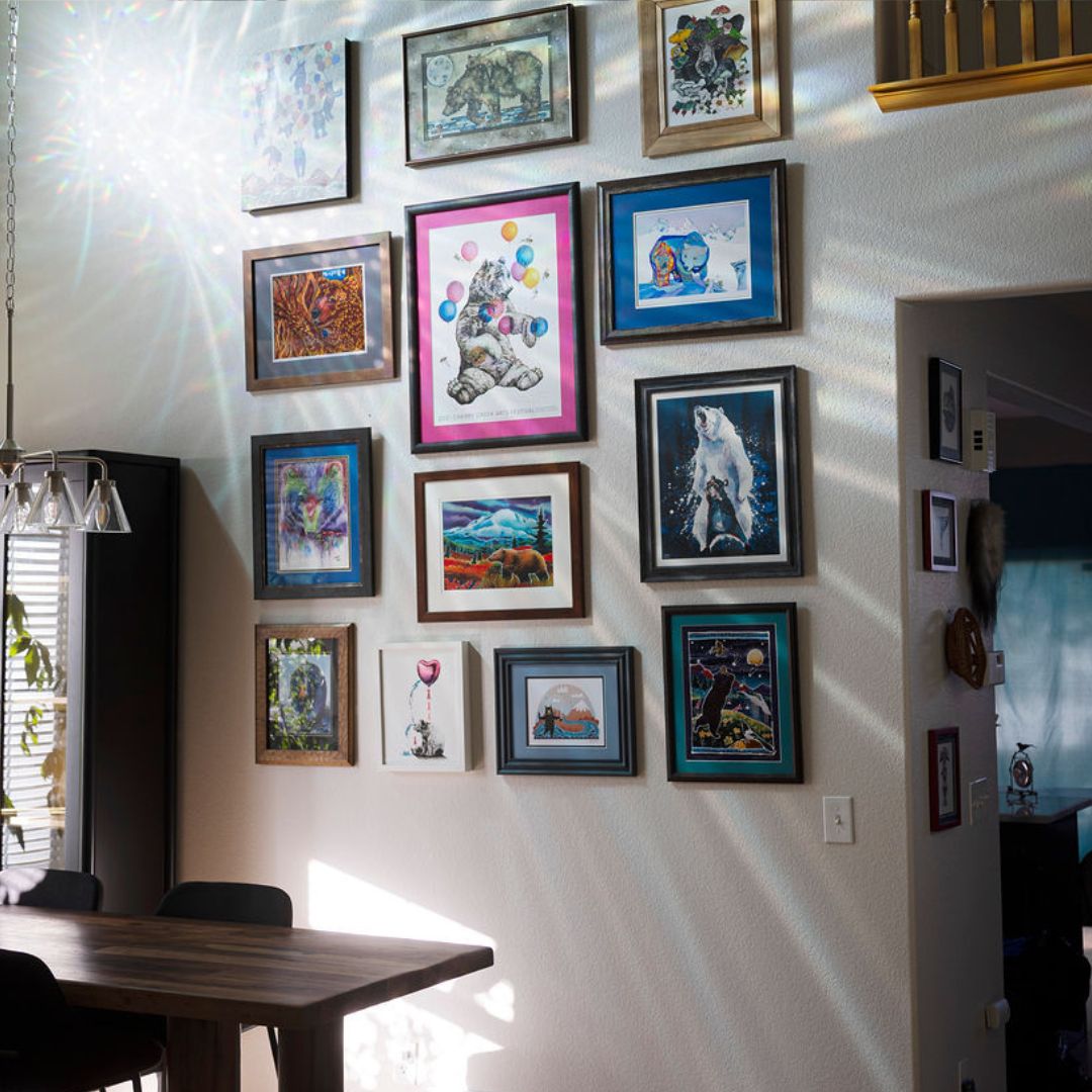 A gallery wall in a dining room featuring multiple framed art prints and paintings in various colors and styles, with sunlight casting dramatic shadows on the wall.