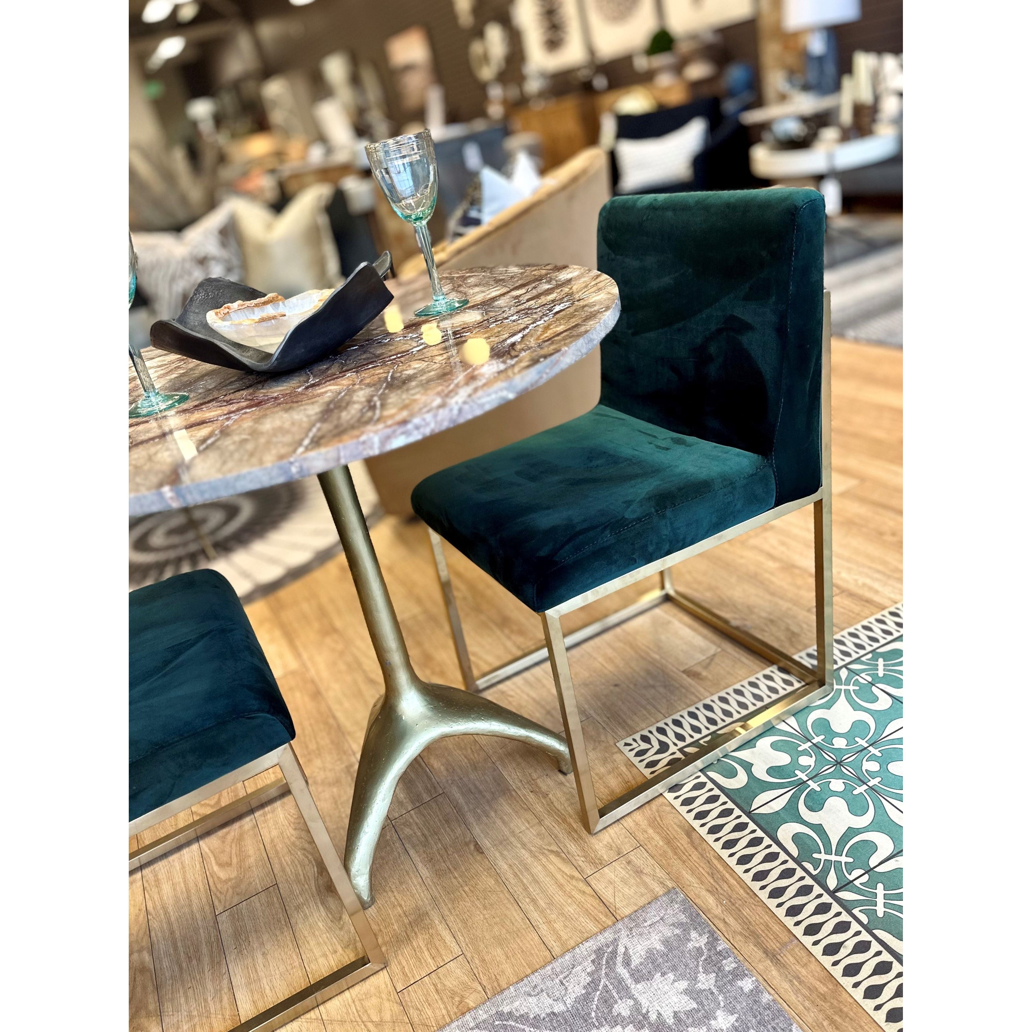 A modern dining space exudes glamour with a round table featuring a marble-patterned top and gold metal base. The Green Velvet Dining Chair, with its gold frame, pairs elegantly alongside a wine glass, black plate, and wooden accents against wood flooring and patterned tiles.
