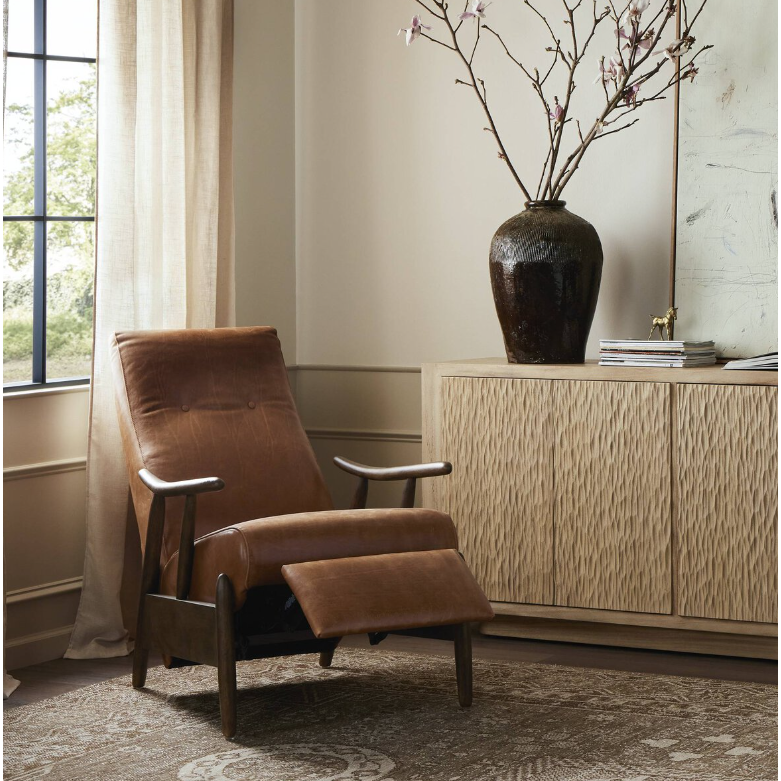 A cozy corner flaunts the Harrison Leather Recliner in rich brown, featuring wooden armrests, positioned beside a beige-patterned sideboard. A large dark vase with branches sits atop the sideboard. Natural light filters through a sheer curtain by the window, illuminating the carpeted floor.