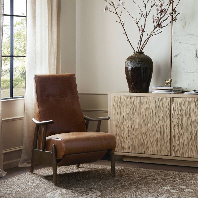 A cozy living space featuring the Harrison Leather Recliner, which boasts brown leather and wooden armrests, sits by a large window with beige curtains. On the right, there’s a light-colored textured cabinet topped with a large dark vase holding branches with pink flowers. The floor is adorned with a patterned rug.