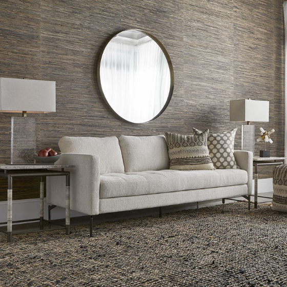 In a contemporary living room, a round mirror adorns the textured brown wall. Below, the Victory Chenille Sofa in beige, accented with patterned cushions, sits between two square side tables. One holds a decorative bowl of apples; the other displays a white flower arrangement. A woven rug completes the look.