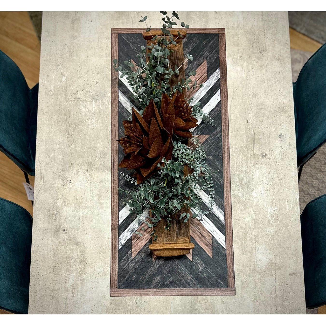 A rectangular wooden table with a black, white, and brown geometric runner features outdoor decor—a central arrangement with faux greenery, eucalyptus, large rust-colored leaves, and the Rust Aster Metal Flower. Four dark teal chairs surround the table.