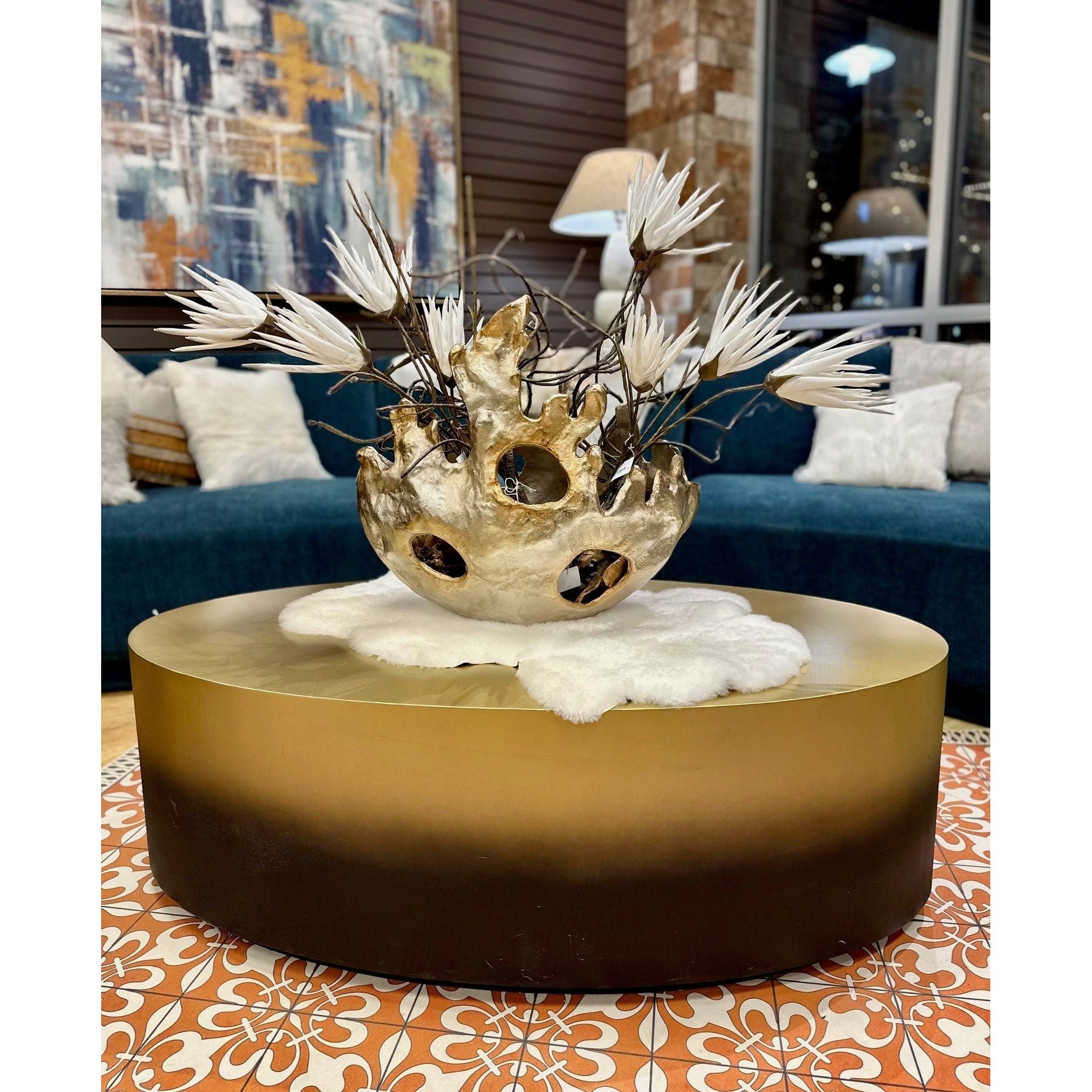 A Dolce Coffee Table in ambre black and gold showcases a textured ceramic vase of spiky white flowers and black branches, paired with a dark teal sofa with beige pillows. An abstract painting adds modern elegance, while city lights twinkle beyond the windows.