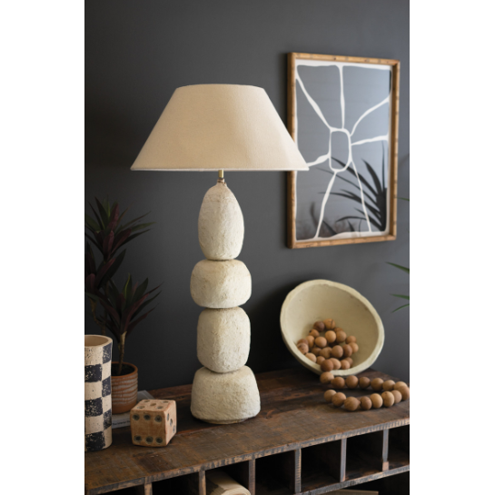 A Quad Paper Mache Lamp with a textured stone base and natural fabric shade rests on wood. Nearby, wooden balls fill a bowl, flanked by a checkered vase and patterned cube. An abstract painting enhances the dark gray wall, complemented by partially visible plant leaves for an artistic vibe.