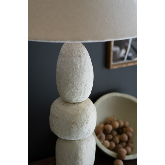 A close-up of the Quad Paper Mache Lamp, featuring a textured beige base of stacked rounded forms and a neutral fabric shade. In the background, a bowl of wooden balls rests near a dark wall and framed picture.