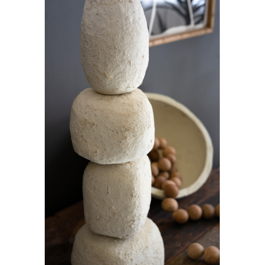 A vertical stack of four large, rough-textured, beige forms makes up the Quad Paper Mache Lamp, placed on a wooden surface. Behind it sit a beige bowl of round brown nuts, some scattered nearby, with a mirror on the gray wall.