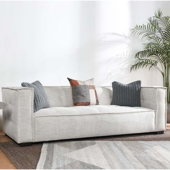 The Princeton Sofa in beige features modern button tufting and comes with three patterned cushions in gray, brown, and white. It sits on a geometric-patterned rug in gray and brown tones, with a tall green plant nearby as sunlight creates soft wall shadows.