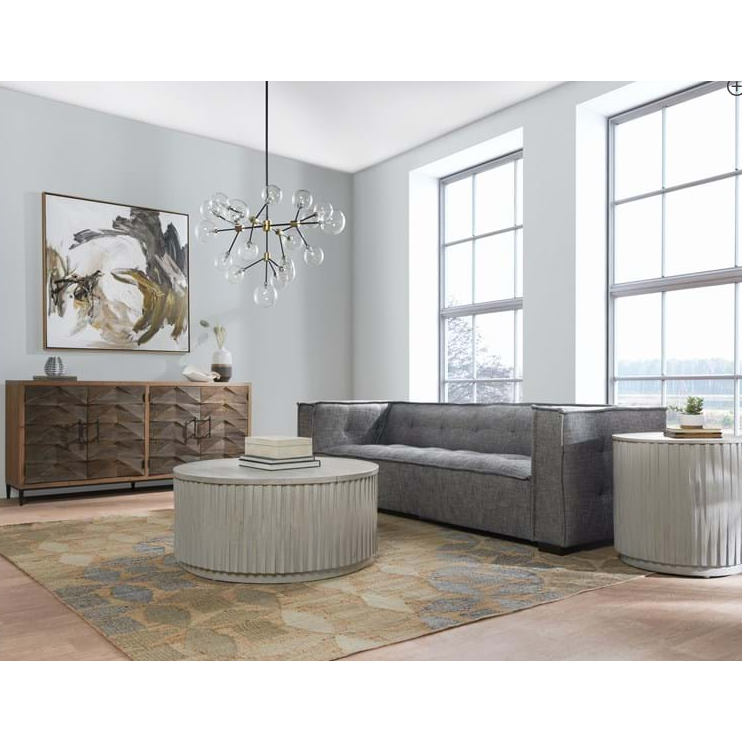 A modern living room features the Princeton Sofa in charcoal with tufted details, complemented by abstract art and a wooden sideboard. A round, textured coffee table and matching side table sit on a patterned rug. Large windows allow natural light in, while a glass orb chandelier hangs above.