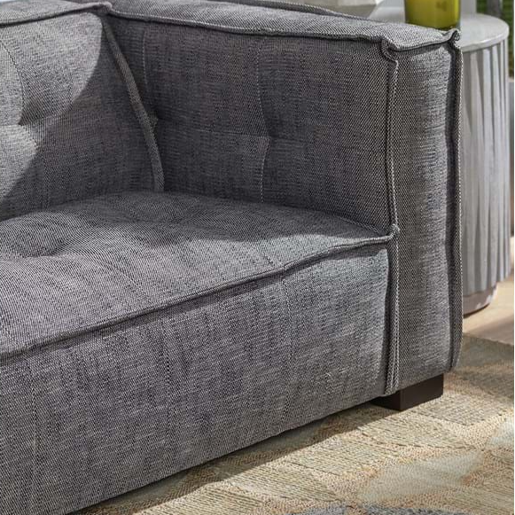 Close-up of the Princeton Sofa in charcoal, showcasing tufted detailing and cushioned seats in durable polyester with visible stitching and textured upholstery. Partially visible are a side table with vertical grooves and a green decor piece in the background.