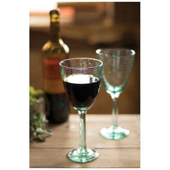 Two Recycled Glass Wine Glasses with a green tint rest on a wooden table, one brimming with red wine while the other remains unfilled. In the background, a blurred wine bottle and gentle, warm lighting enhance the cozy atmosphere.