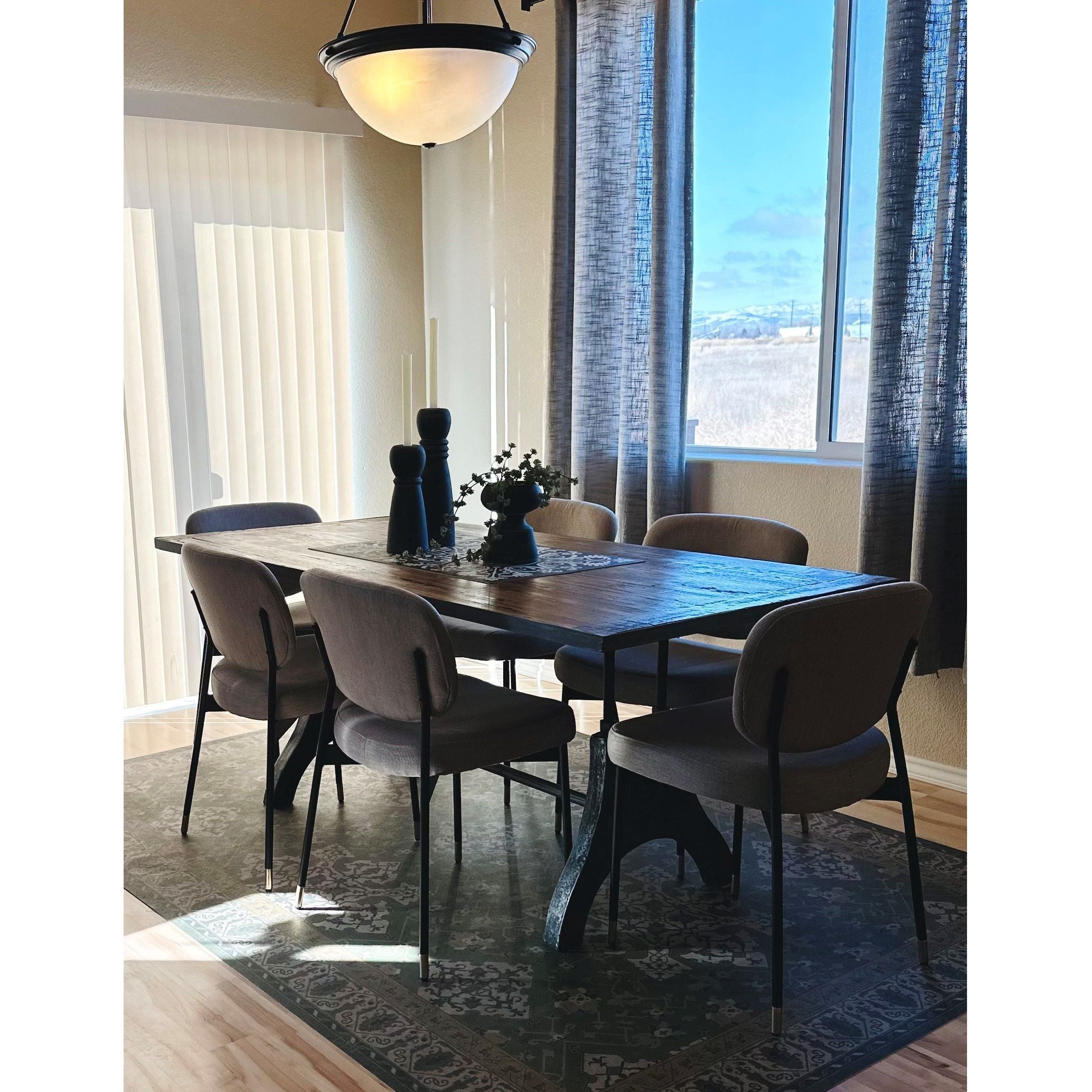 A dining room showcases a wooden table with a sleek black metal frame, encircled by six Ridge Dining Chairs. The tabletop is embellished with a decorative centerpiece and elegant black candlesticks, adding an air of luxury. Natural light streams through a large window dressed with sheer curtains, illuminating the patterned rug below.