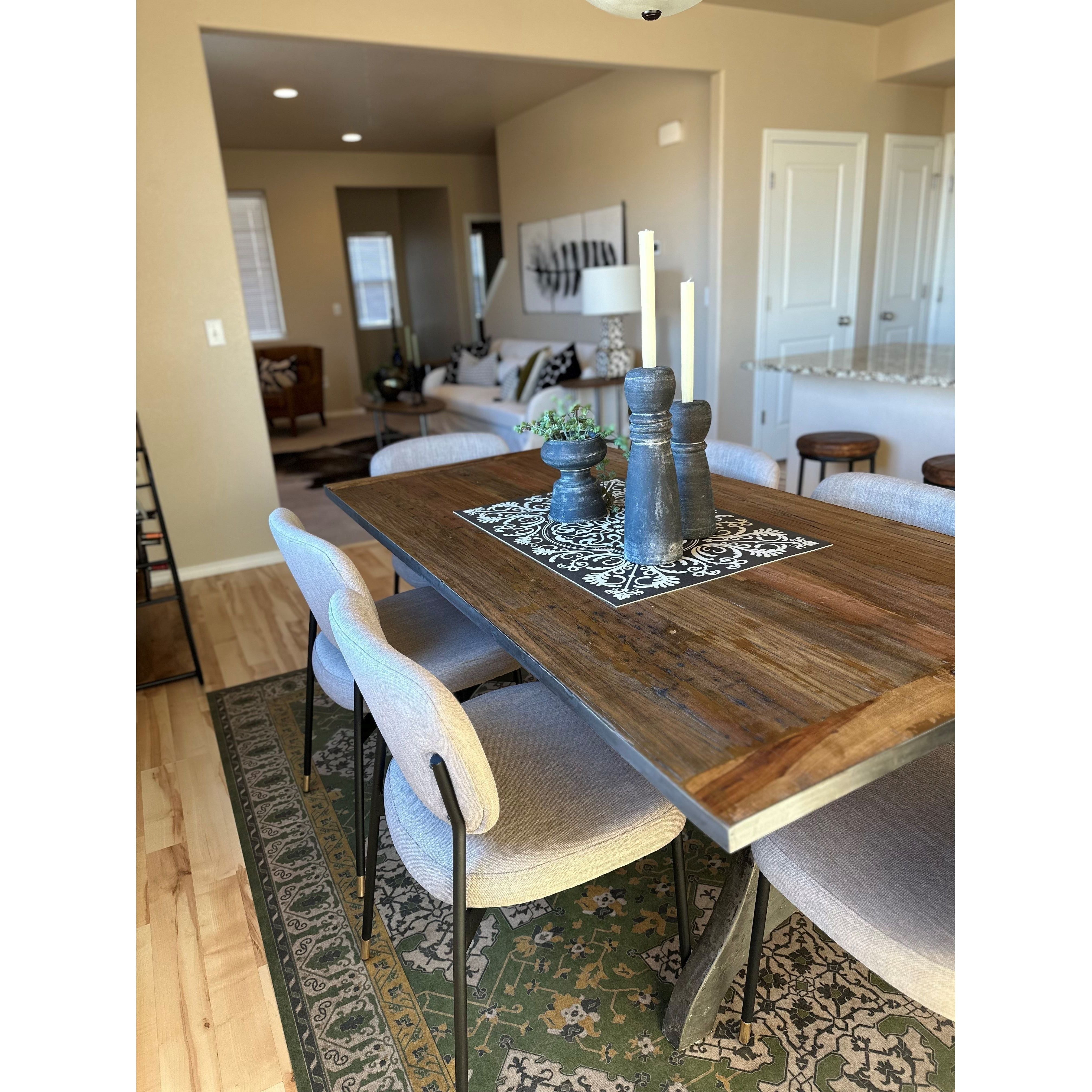 The dining room boasts a wooden table adorned with a decorative runner, black candle holders, and a small plant. Surrounding the table are four Ridge Dining Chairs, adding a luxurious touch to the setting. In the background, there is a cozy living area featuring a sofa, artwork, and abundant natural light streaming through the windows.