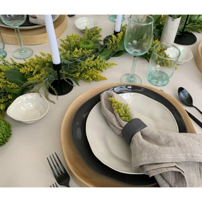 A table setting featuring layered plates atop a Wood Charger and a sustainably sourced, beige cloth napkin adorned with greenery in a black napkin ring, paired with black cutlery. The centerpiece showcases a garland of green leaves and yellow flowers. Black candle holders with tall white candles and elegant glassware complete the look.
