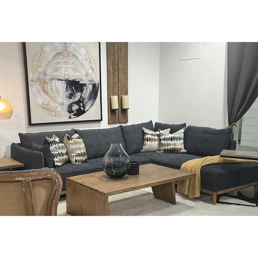 A contemporary living room features a gray Nathanael Grey Blue Sectional with decorative black and white pillows, a wooden coffee table, a round abstract painting above the sofa, complemented by a cozy gray curtain and a soft white rug.