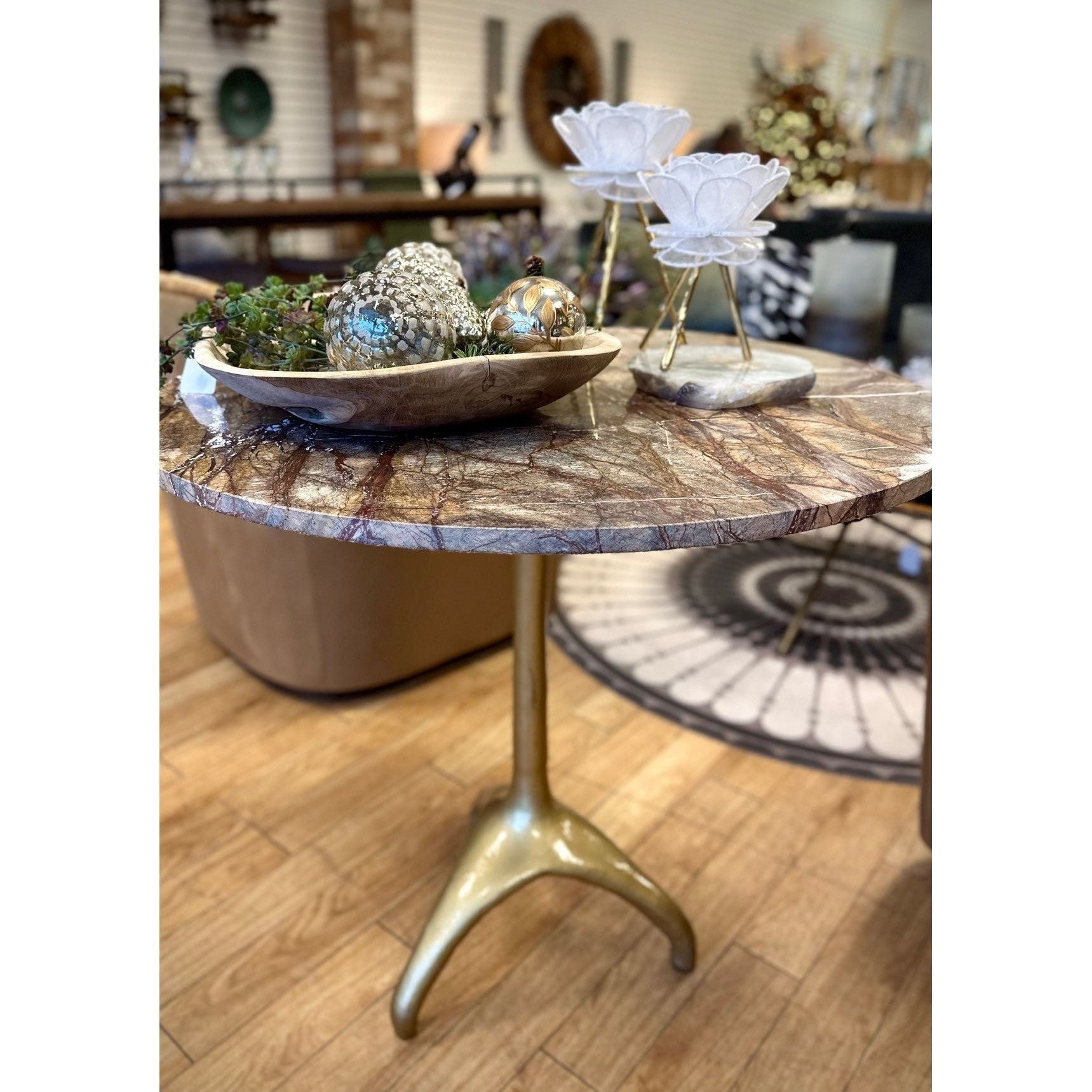 The Sierra Café Table stands in a showroom with an antique-finished Bidasar Brown Marble top and a brass-finished cast-iron base. On the table, there's a wooden bowl filled with decorative items, including dried flowers and metallic balls. Two white flower-shaped objects on gold stands are placed next to the bowl. The background shows furniture and decor items.
