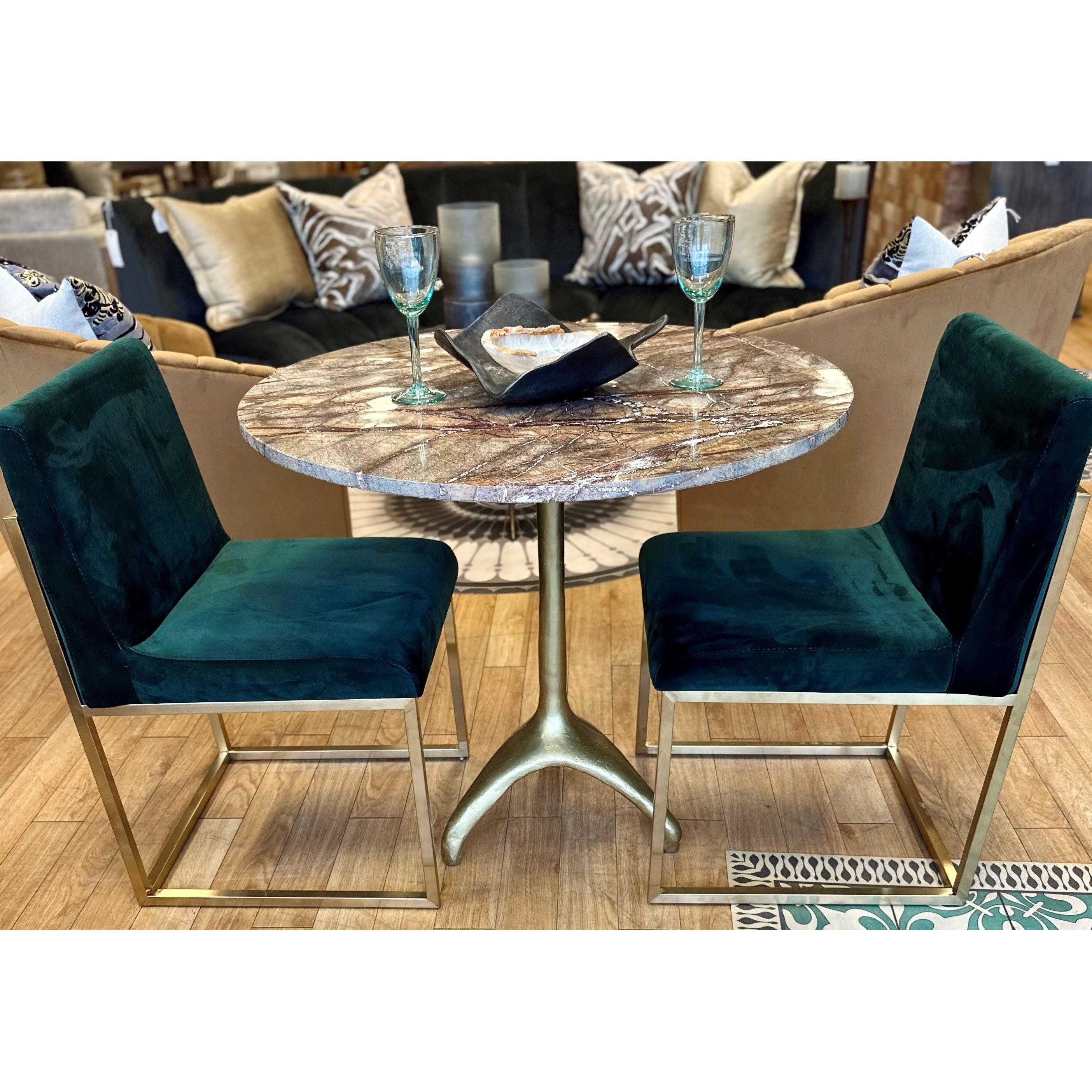A cozy dining setup featuring a round marble table with a gold pedestal base, surrounded by two modern glamour Green Velvet Dining Chair. The table is set with two teal wine glasses and a small snack bowl on a black napkin. A beige sofa with patterned cushions is visible in the background.