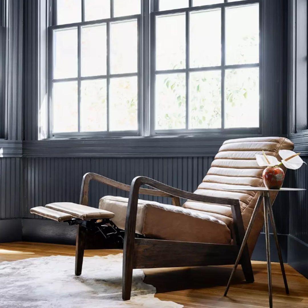 The Terra Leather Recliner, Taupe, with top-grain leather and dark wooden arms, sits by large black-framed windows. Sunlight highlights a cowhide rug and a small table topped with a decorative vase and books.