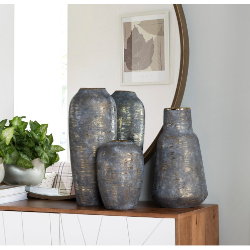 Four Triad Vases with a blue-gray and gold accent finish are displayed on a modern wooden sideboard. Behind them are a green potted plant, a round mirror, and framed botanical leaf artwork.
