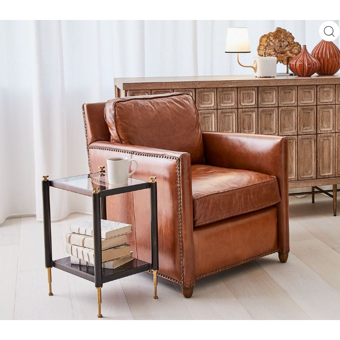 In a cozy living room, a Winston Leather Club Chair crafted in brown full-grain leather with antique bronze nailhead trim adds elegance. Next to it, a sleek glass and metal side table supports a white mug and stacked books. A wooden sideboard with decorative vases and an illuminated lamp enhances the background, while white curtains diffuse natural light.