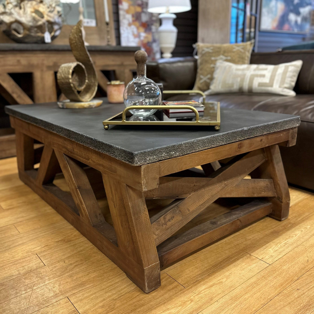 The Alderbrook Wood and Stone Coffee Table, with a thick stone top and geometric wood base, sits on a light wood floor. A decorative tray with a glass decanter and metal accents rests on it, with a brown sofa and decor in the background.