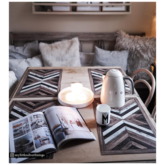A cozy table setup featuring Wood Art Vinyl Placemats with geometric patterns, an open magazine, a monogrammed mug, a SMEG kettle, three lit candles on a tray, plus plush cushions and a fluffy throw for extra comfort.
