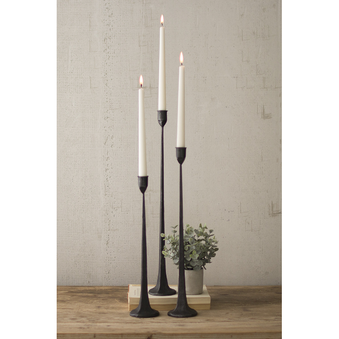 Three lit white taper candles are placed in sleek, tall William Cast Iron Candlesticks of varying heights, standing on a wooden surface against a light-textured wall. A small potted plant sits nearby atop a stack of two books, adding a touch of greenery to the sophisticated home decor.