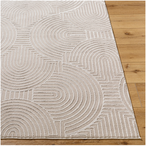 In the image, there's a corner of the Zen Circles Machine Washable Rug, showcasing its durable beige construction with a modern geometric pattern that includes concentric arcs and lines. The medium pile colors stand out against a light wooden floor, providing texture with its raised sections in contrast to the flat background.