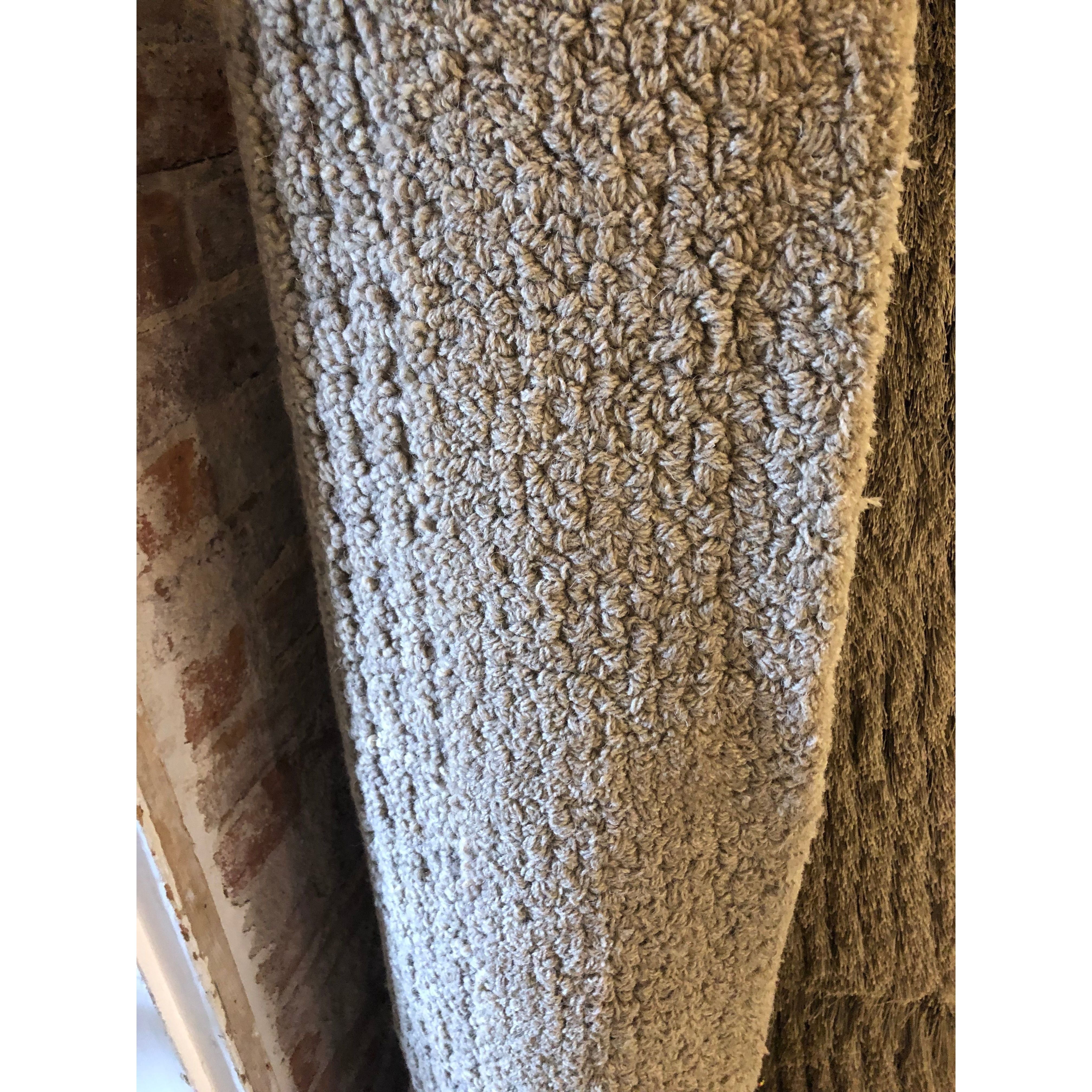 A close-up of the Dejon Wool Rug, rolled vertically beside a rough brick wall. Its beige color, looped fibers, and thick woven pattern showcase the rug’s plush texture and dense construction.