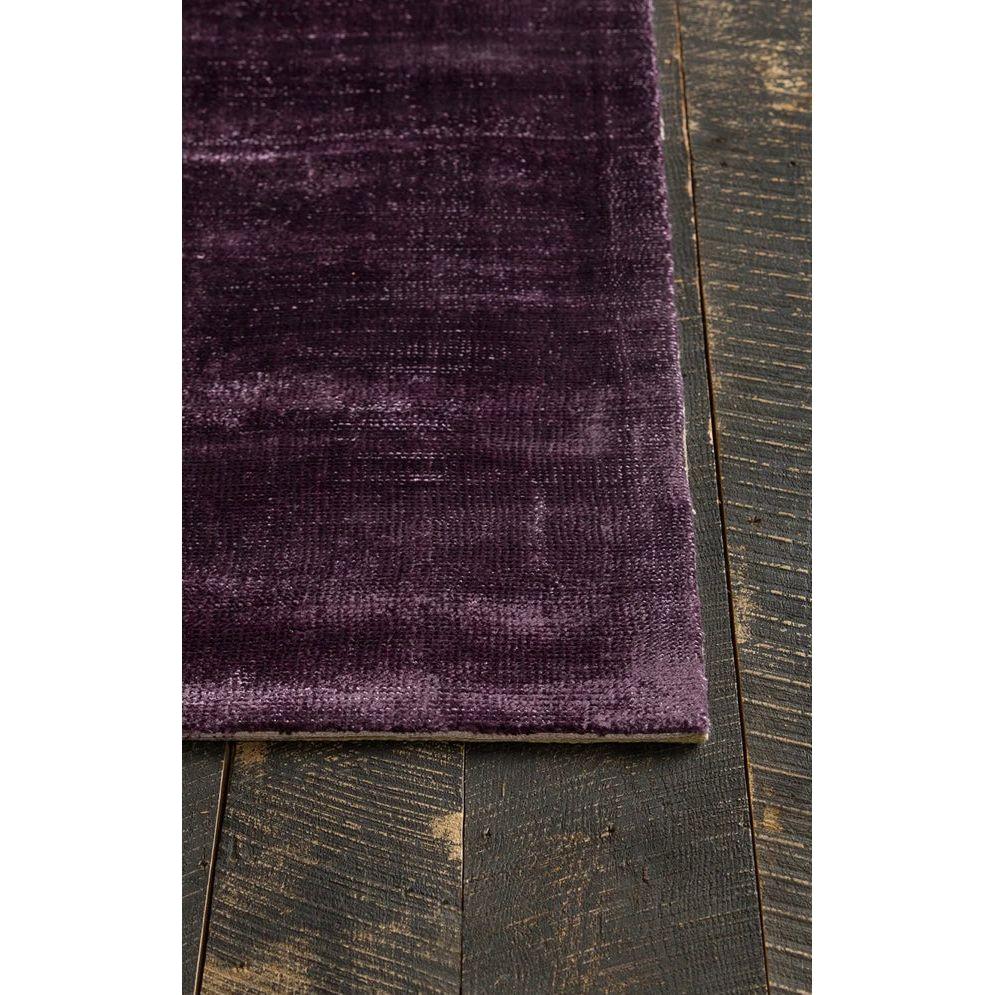 A close-up of the Sopris Art Silk Rug in deep purple velvet rests on dark, textured wood floorboards, its smooth, luxurious fabric contrasting with the rustic, weathered grain beneath.
