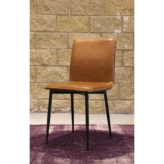 The Lucy Leather Dining Chair, featuring a high backrest in tan leather and slim black metal legs, stands on a purple rug. Positioned against a textured brick wall, its sleek black iron frame highlights the contrast between warm leather, dark metal, and the vibrant background.