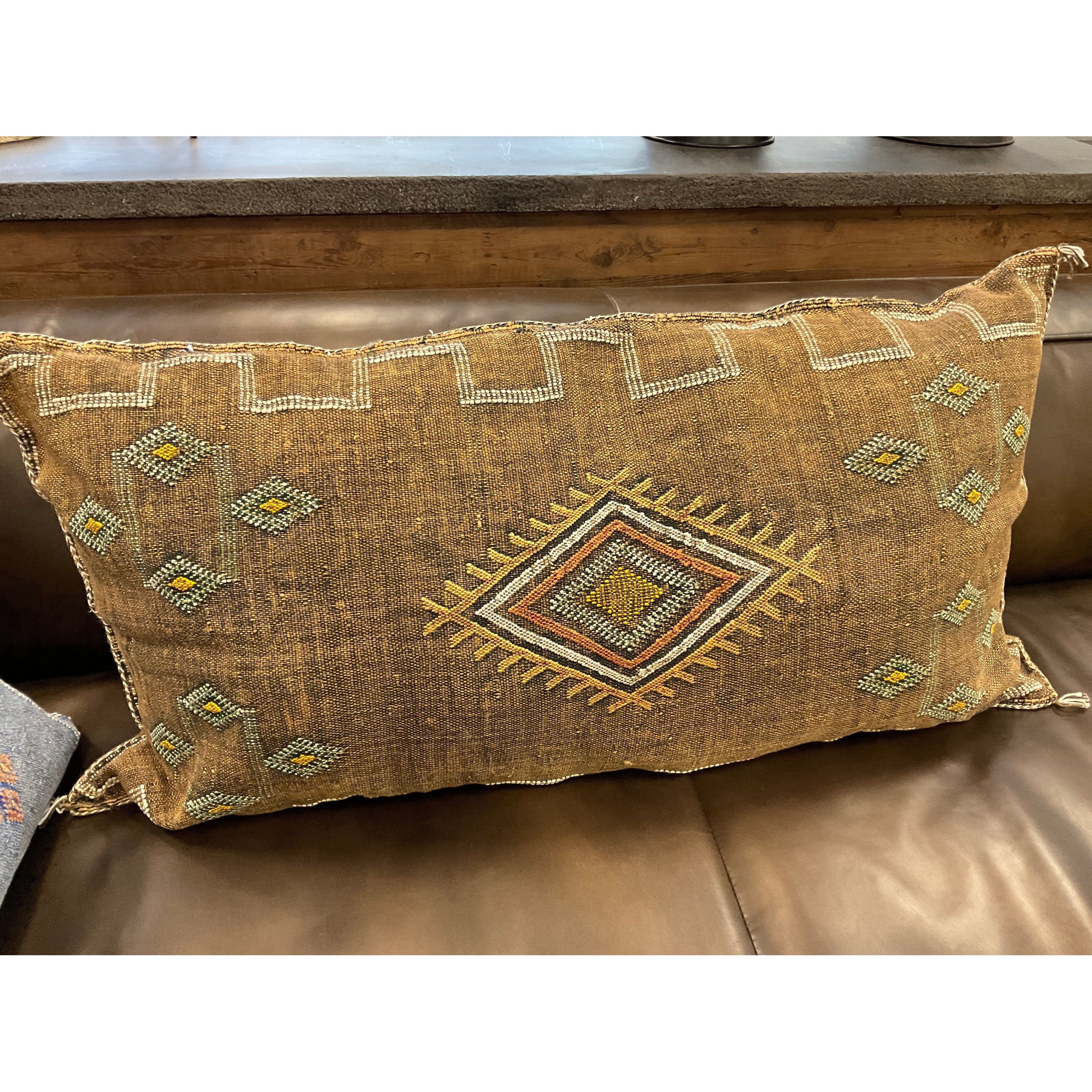 The Earth Cactus Fiber Throw Pillow flaunts a tribal design with intricate weaving and a central diamond pattern in yellow, red, and blue. It's set on a leather couch against a brown backdrop, complemented by wooden ledges and decor items.