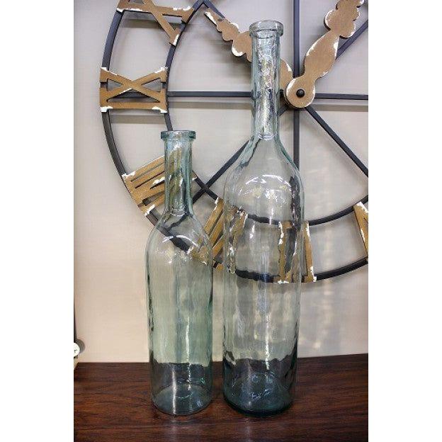 Two Glass Bottle Vases of different heights, one measuring 7"W x 30"H and the other 8"W x 40"H, are positioned in front of a large, partially visible wall clock with Roman numerals and gold-colored clock hands. The transparent vases have a slight green tint and elongated necks, placed on a wooden surface.