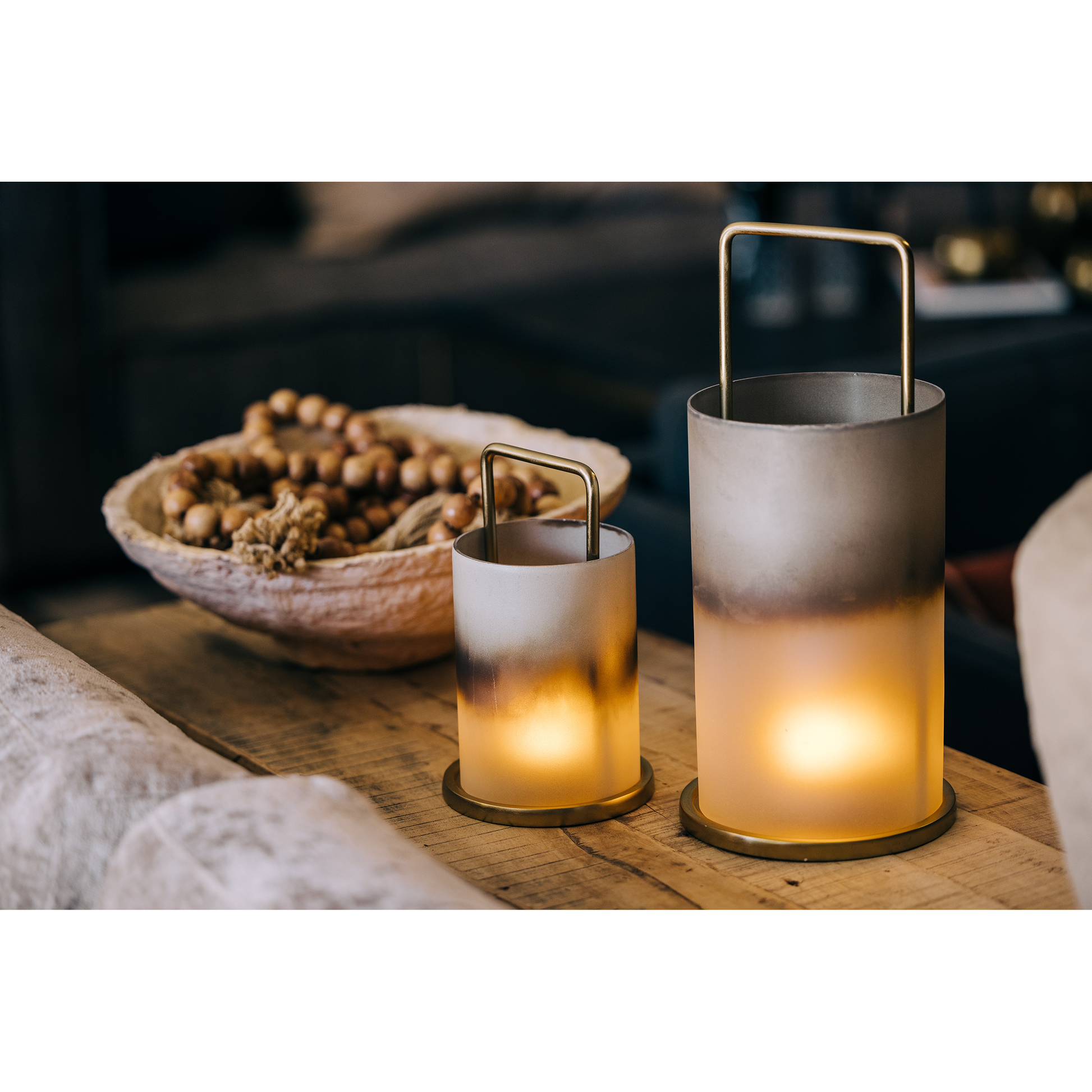 Two cylindrical candle holders with metal handles, one large and one small, emit a warm glow on a wooden table. Enhanced by Rechargeable LED Candles with realistic flame effects and 20+ hour run times, the cozy ambiance is complemented by a bowl filled with nuts in the background.